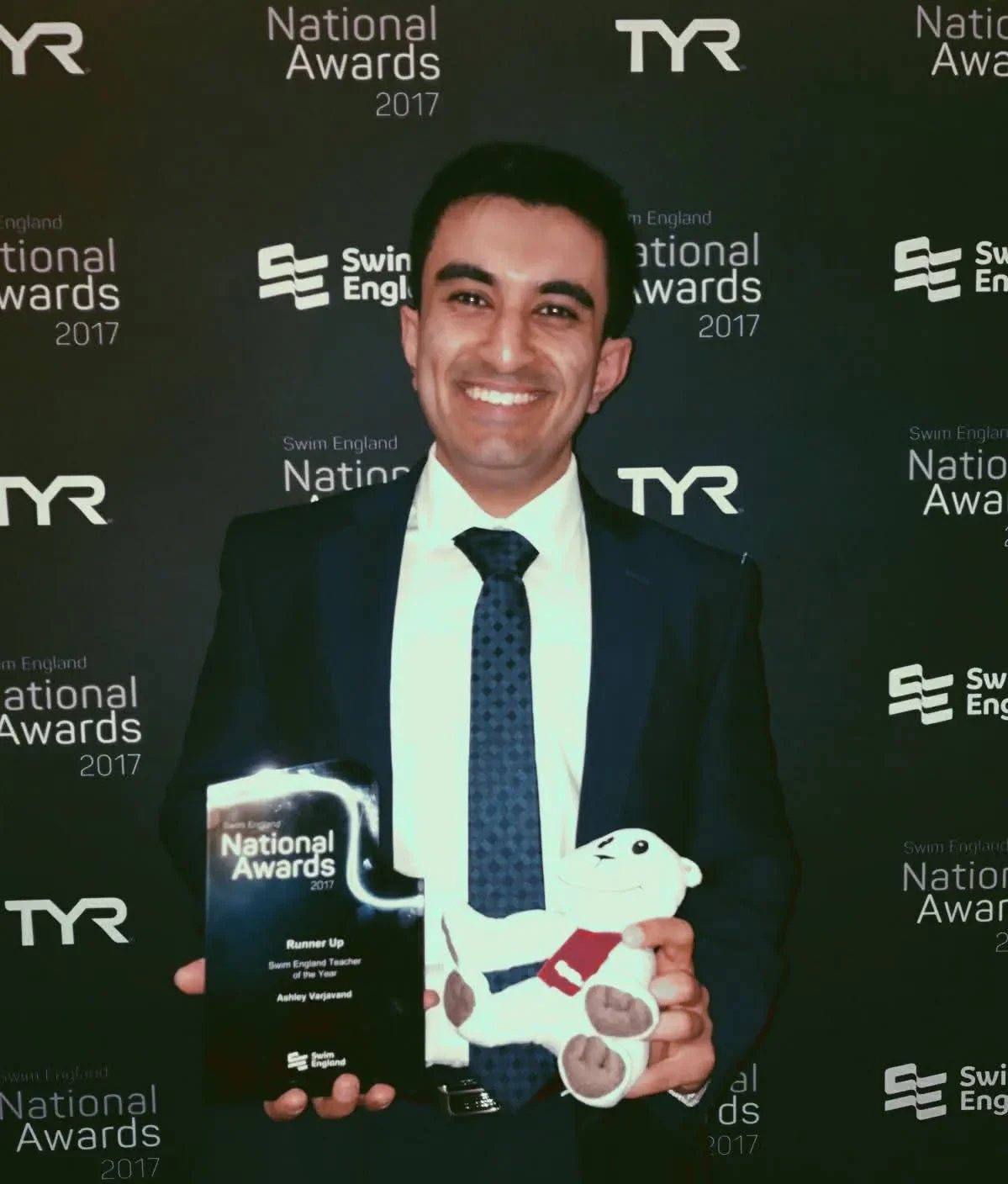 Ashley Varjavand holding the Swim England National Awards 2017 runner-up trophy in front of the branded event backdrop.